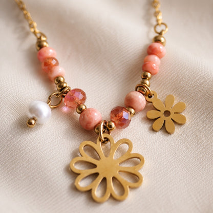 Nahaufnahme goldene Flower Power Halskette auf cremefarbenem Seidenstoff – Detailansicht rosa Rhodonit-Perlen, facettierte Kristalle, Süßwasserperle und zwei vergoldete Blumen-Charms aus Edelstahl
