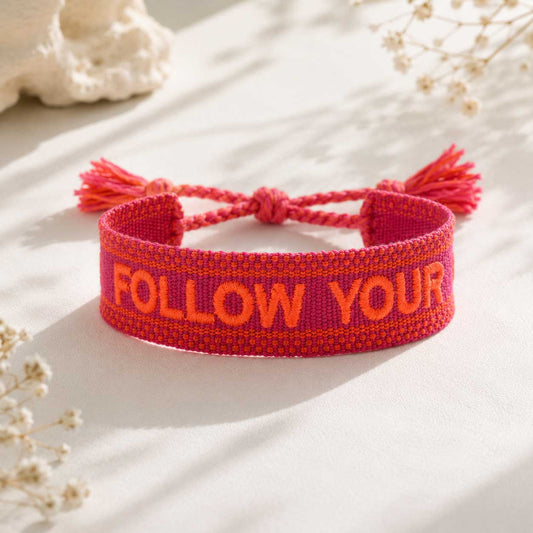 Follow Your Heart Statement Armband – pinkes Webarmband von Saltyblush mit orangefarbener Stickerei und Quasten, verstellbar, Flatlay auf weißem Stoff mit Trockenblumen
