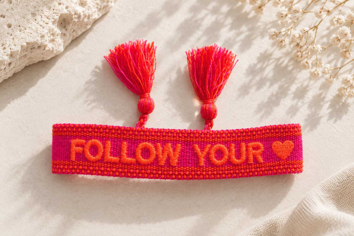 Follow Your Heart Armband Flatlay – pinkes Webarmband von Saltyblush mit Aufschrift "Follow Your Heart" und Herz-Motiv, rote Quasten, Draufsicht auf weißem Stoff
