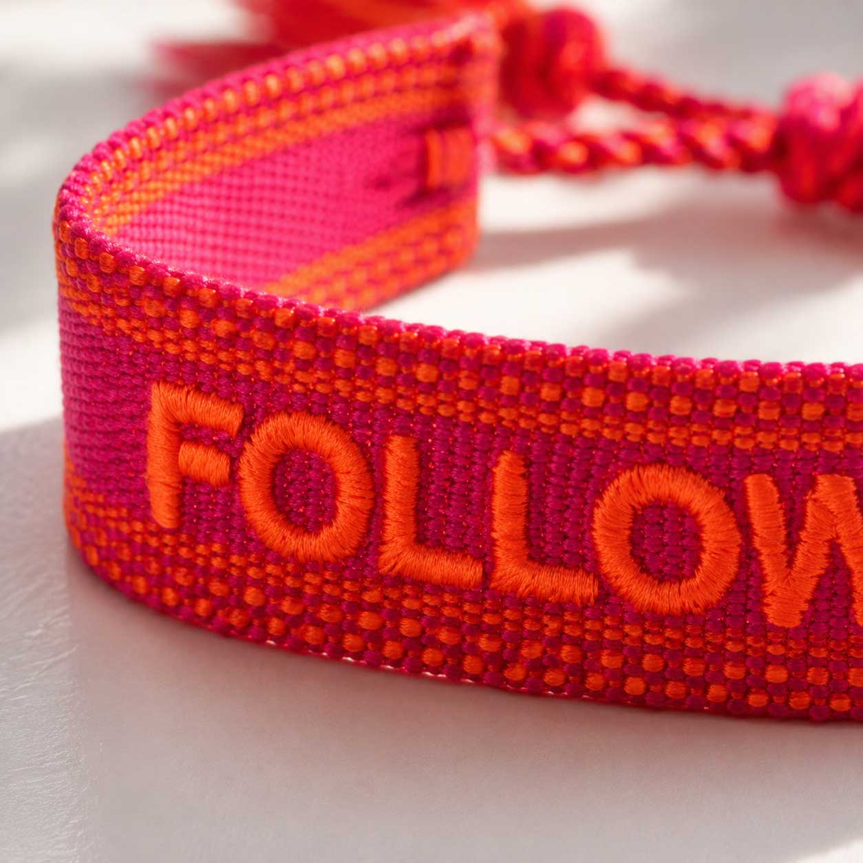 Nahaufnahme Stickerei – Follow Your Heart Armband von Saltyblush, orangefarbene Buchstaben auf pinkfarbenem Webband, strukturierte Textildetails im Sonnenlicht
