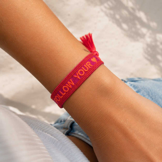 Follow Your Heart Armband am Handgelenk – pinkes Webarmband von Saltyblush getragen, rote Quaste, sommerlicher Strandlook zu Jeans-Shorts am Meer
