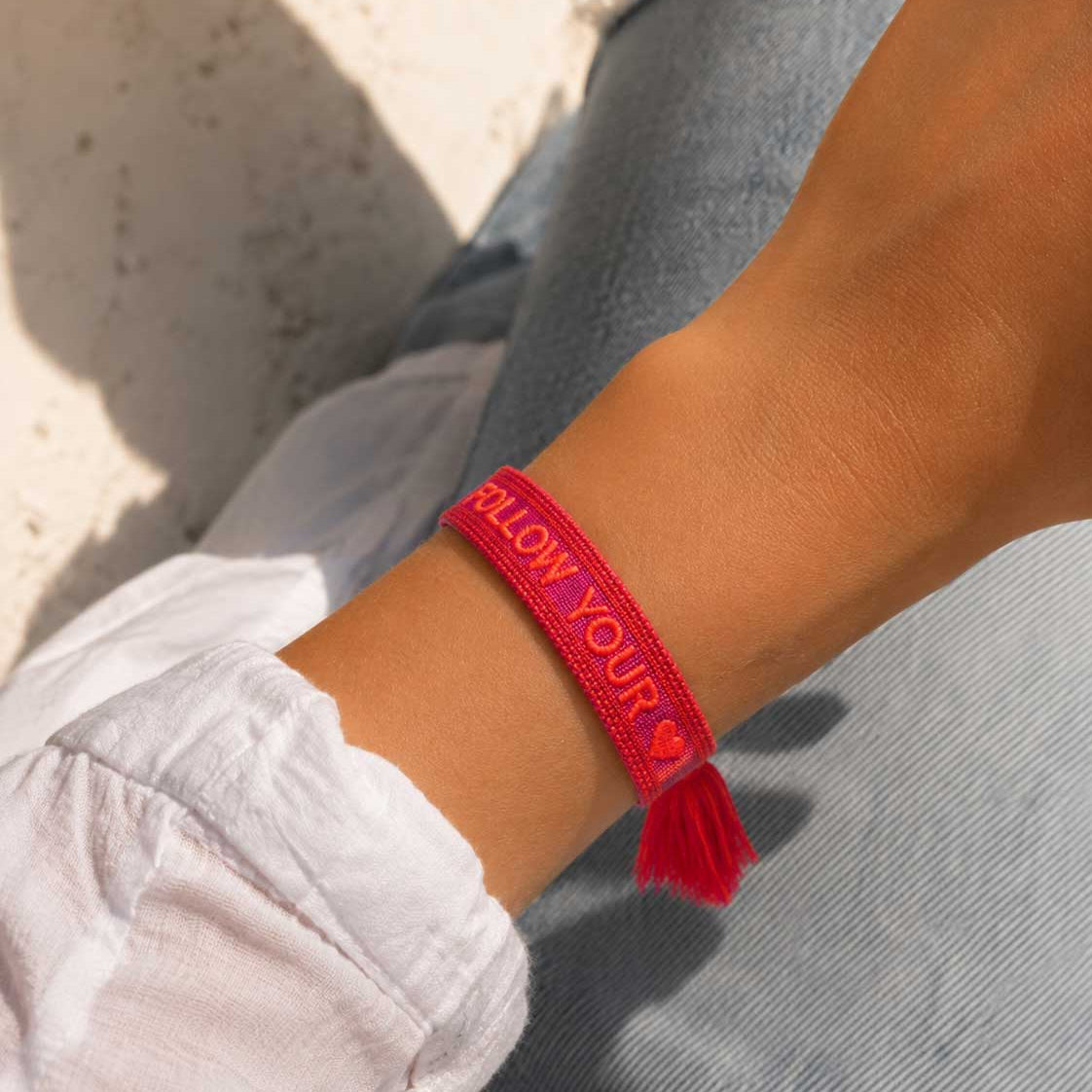 Follow Your Heart Armband Lifestyle – pinkes Webarmband von Saltyblush am Handgelenk, rote Quaste, lässiger Look zu weißem Hemd und Jeans im Sonnenlicht
