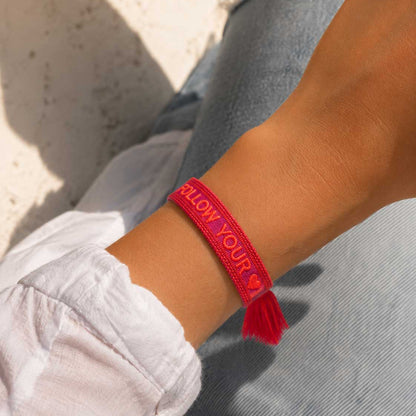 Follow Your Heart Armband Lifestyle – pinkes Webarmband von Saltyblush am Handgelenk, rote Quaste, lässiger Look zu weißem Hemd und Jeans im Sonnenlicht
