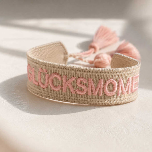 Glücksmomente Statement Armband – beiges Webarmband von Saltyblush mit rosafarbener Stickerei und rosa Quasten, verstellbar, Nahaufnahme im Sonnenlicht

