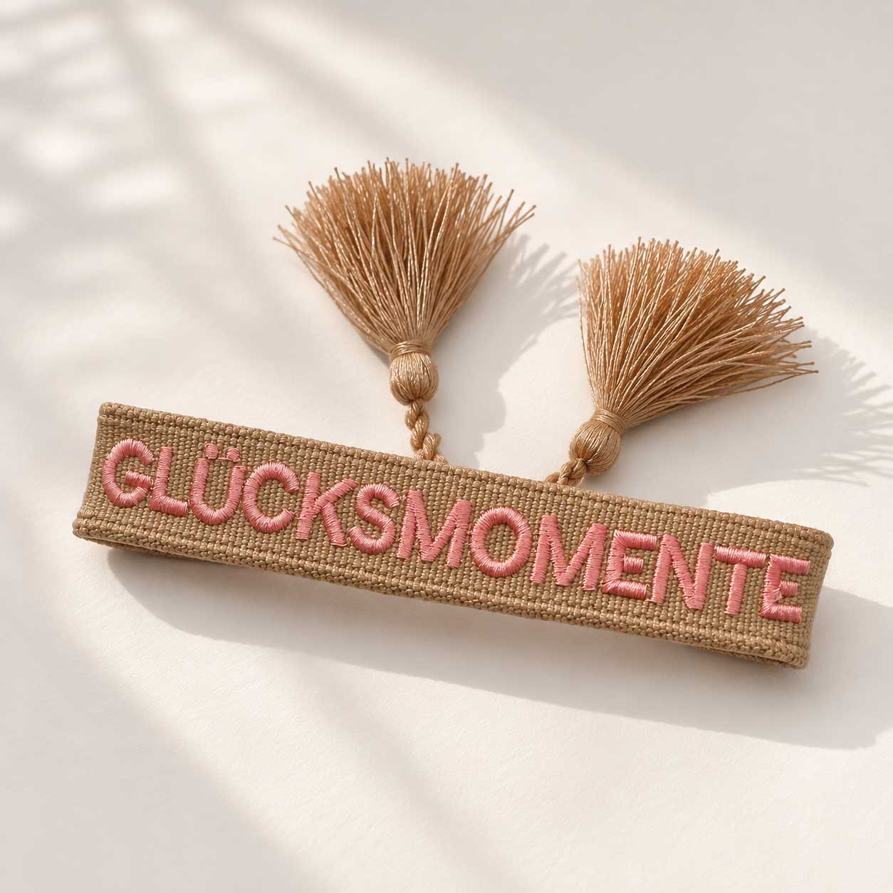 Glücksmomente Armband Flatlay – beiges Webarmband von Saltyblush mit Aufschrift "Glücksmomente" und goldbeigen Quasten, Draufsicht auf weißem Stoff im Sonnenlicht
