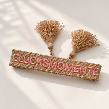 Glücksmomente Armband Flatlay – beiges Webarmband von Saltyblush mit Aufschrift "Glücksmomente" und goldbeigen Quasten, Draufsicht auf weißem Stoff im Sonnenlicht
