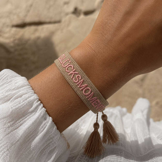Glücksmomente Armband am Handgelenk – beiges Webarmband von Saltyblush getragen, rosafarbene Stickerei mit goldbeigen Quasten, sommerlicher Look zu weißem Oberteil am Strand
