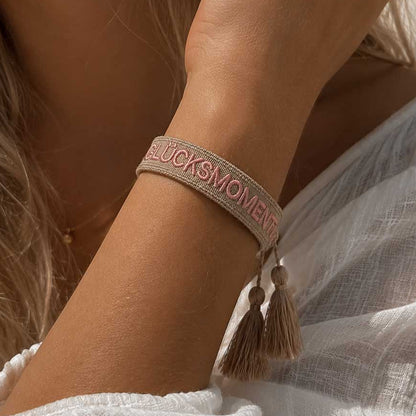 Glücksmomente Armband Lifestyle – beiges Webarmband von Saltyblush am Handgelenk einer Frau mit blondem Haar, goldbeige Quasten, eleganter Look zu weißem Leinenoberteil
