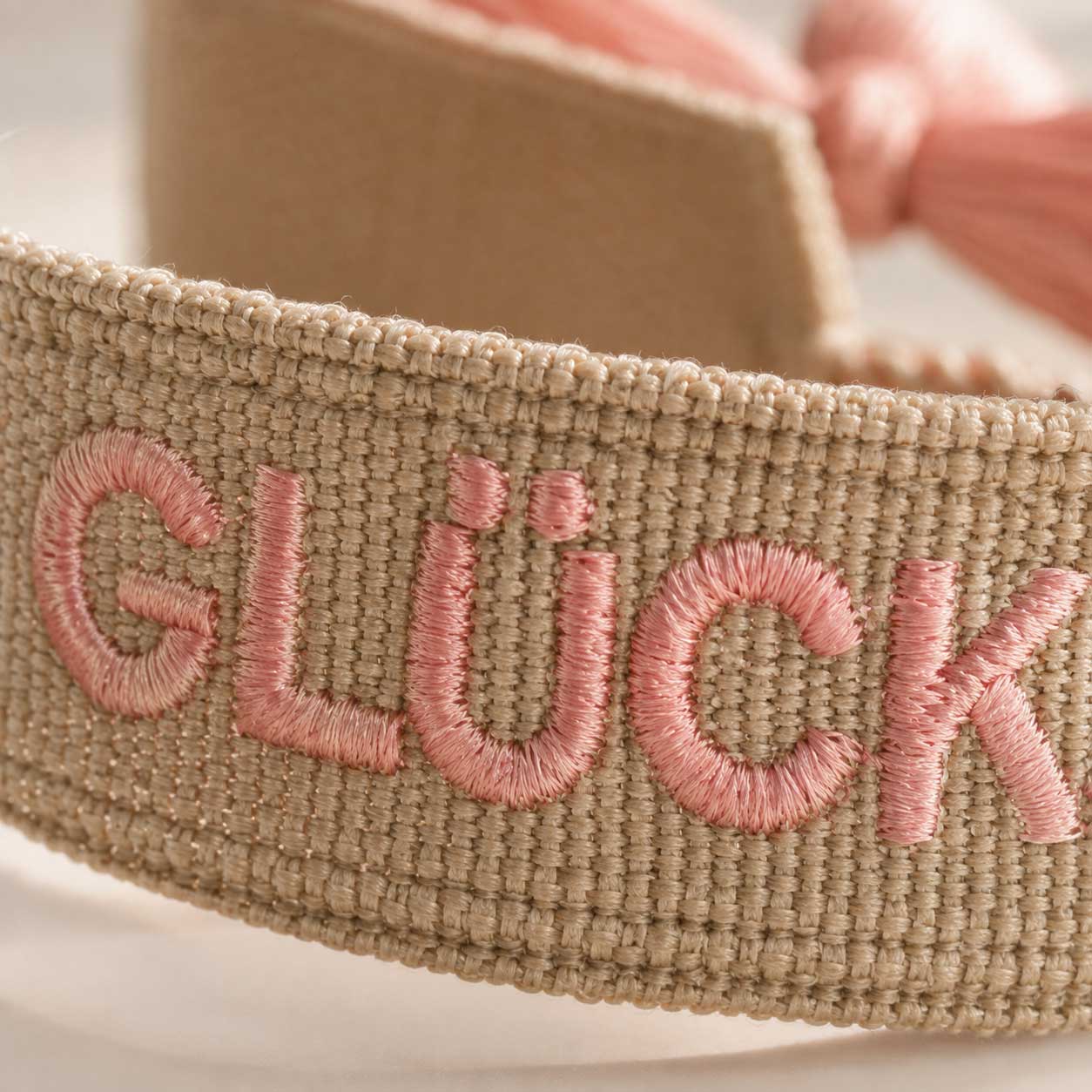 Nahaufnahme Stickerei Glücksmomente – rosafarbene Buchstaben auf beigem Webband von Saltyblush, strukturierte Textildetails, Detailansicht im Sonnenlicht
