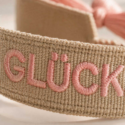 Nahaufnahme Stickerei Glücksmomente – rosafarbene Buchstaben auf beigem Webband von Saltyblush, strukturierte Textildetails, Detailansicht im Sonnenlicht
