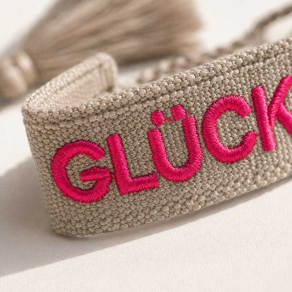 Nahaufnahme gestickter Schriftzug „Glücksmomente" in Pink auf grauem Webarmband – Saltyblush Modeschmuck

