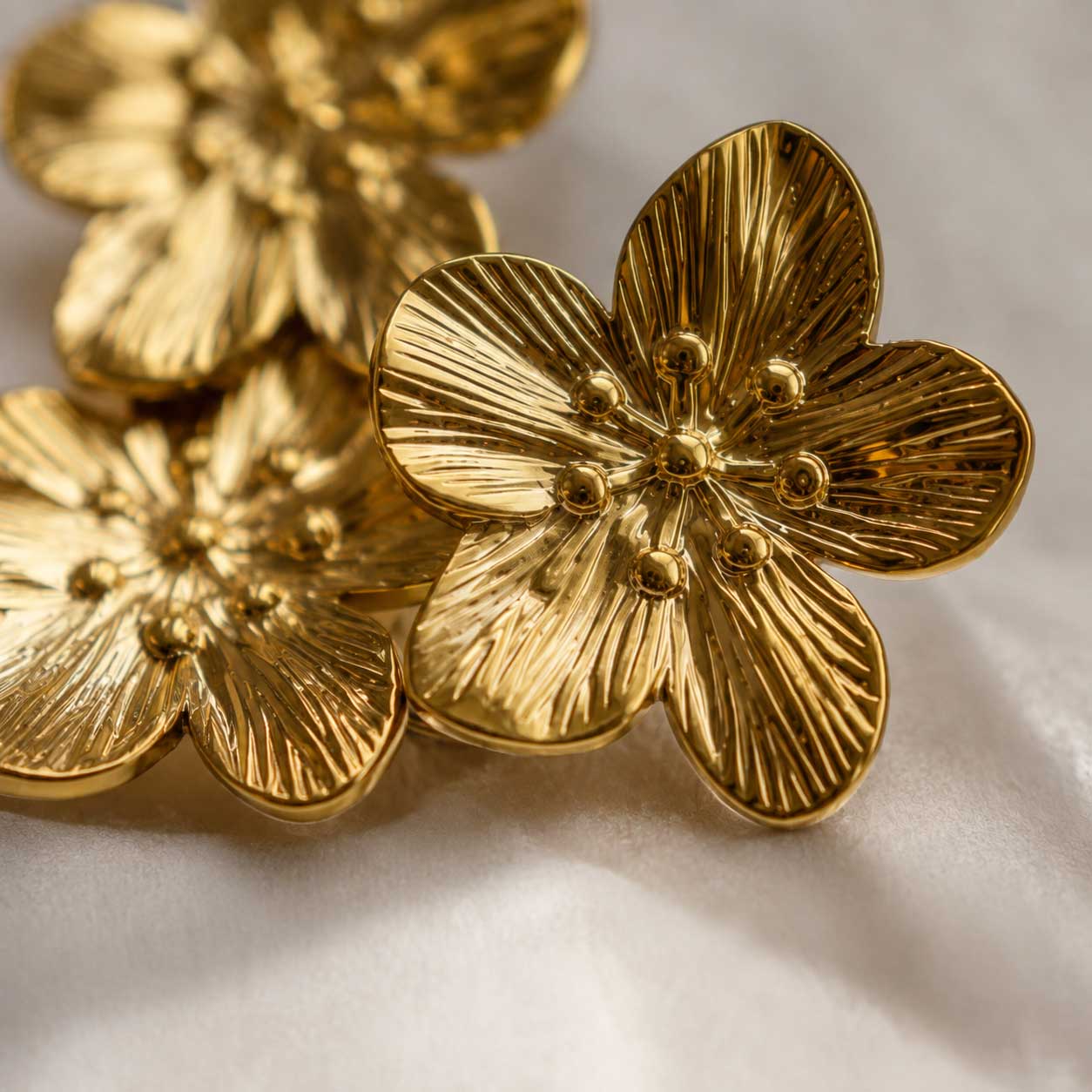 Nahaufnahme der Golden Blossom Ohrringe aus Edelstahl mit detaillierter Blütenstruktur und goldenen Staubgefäßen auf weißem Seidenstoff – Saltyblush
