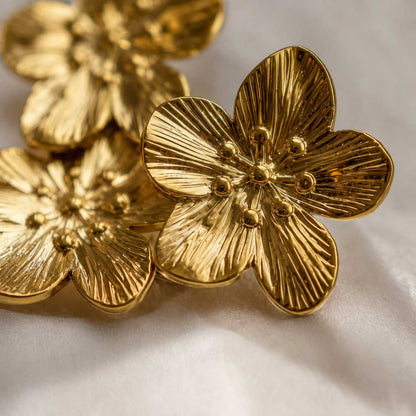 Nahaufnahme der Golden Blossom Ohrringe aus Edelstahl mit detaillierter Blütenstruktur und goldenen Staubgefäßen auf weißem Seidenstoff – Saltyblush
