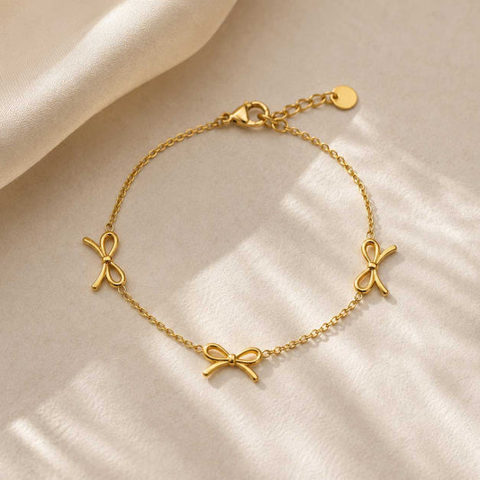 Goldenes Armband mit Schleifenanhängern – Golden Bow Armband von Saltyblush, filigrane Goldkette mit 3 Schleifencharms, Flatlay auf hellem Hintergrund
