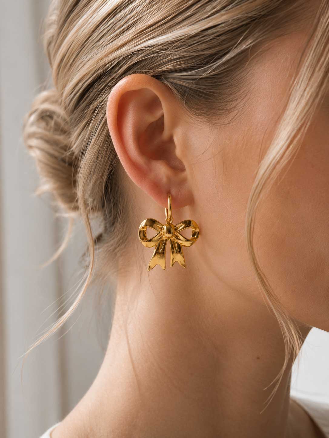 Goldener Schleifen-Ohrring am Ohr getragen, vergoldeter Edelstahl-Ohrhänger mit dreidimensionalem Schleifenanhänger zu blondem Hochsteckhaar und weißem Outfit
