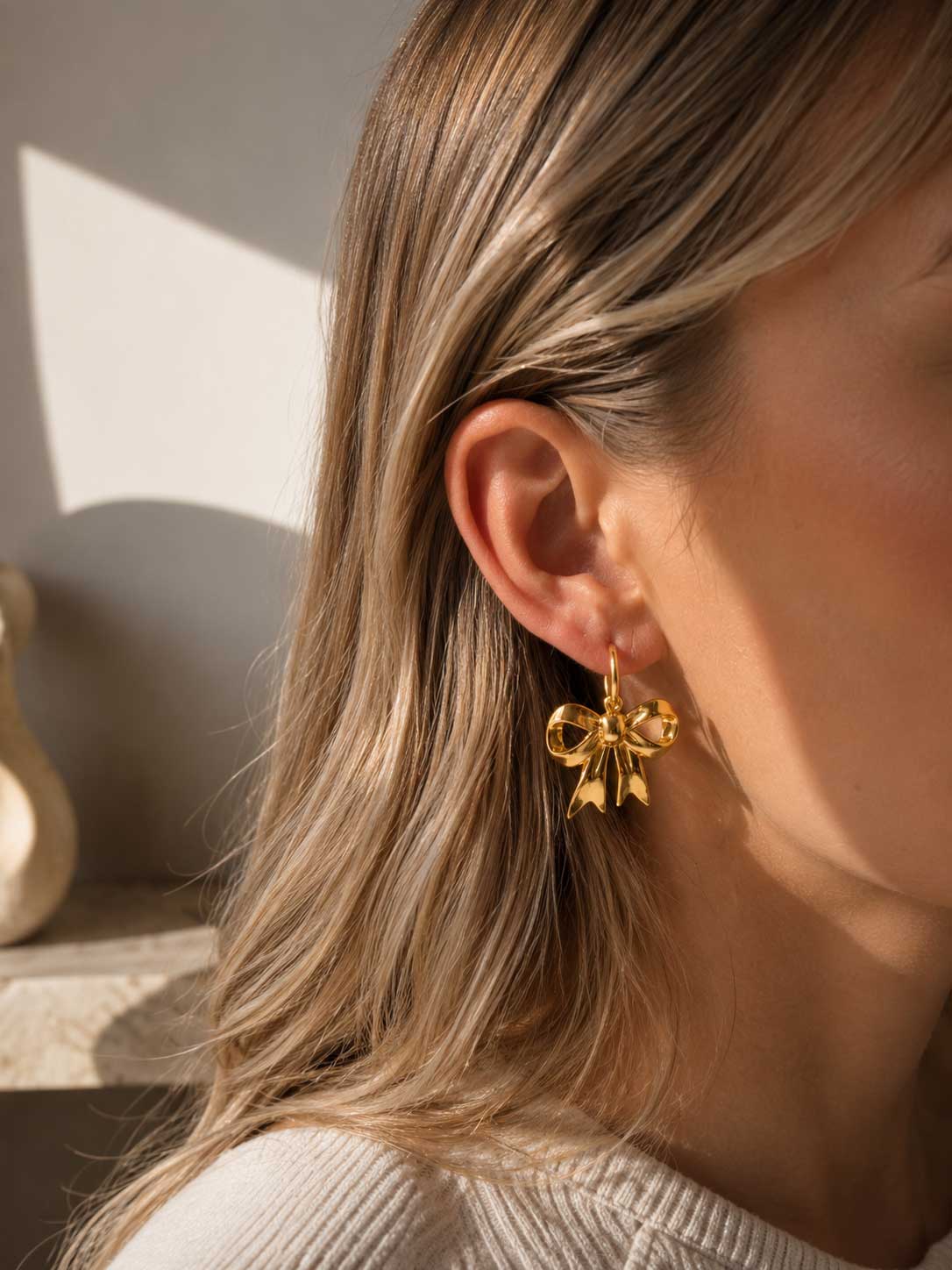 Goldener Schleifen-Ohrring am Ohr getragen im Sonnenlicht, vergoldeter Edelstahl-Ohrhänger mit glänzendem Schleifenanhänger zu offenem blonden Haar und weißem Rippenpullover
