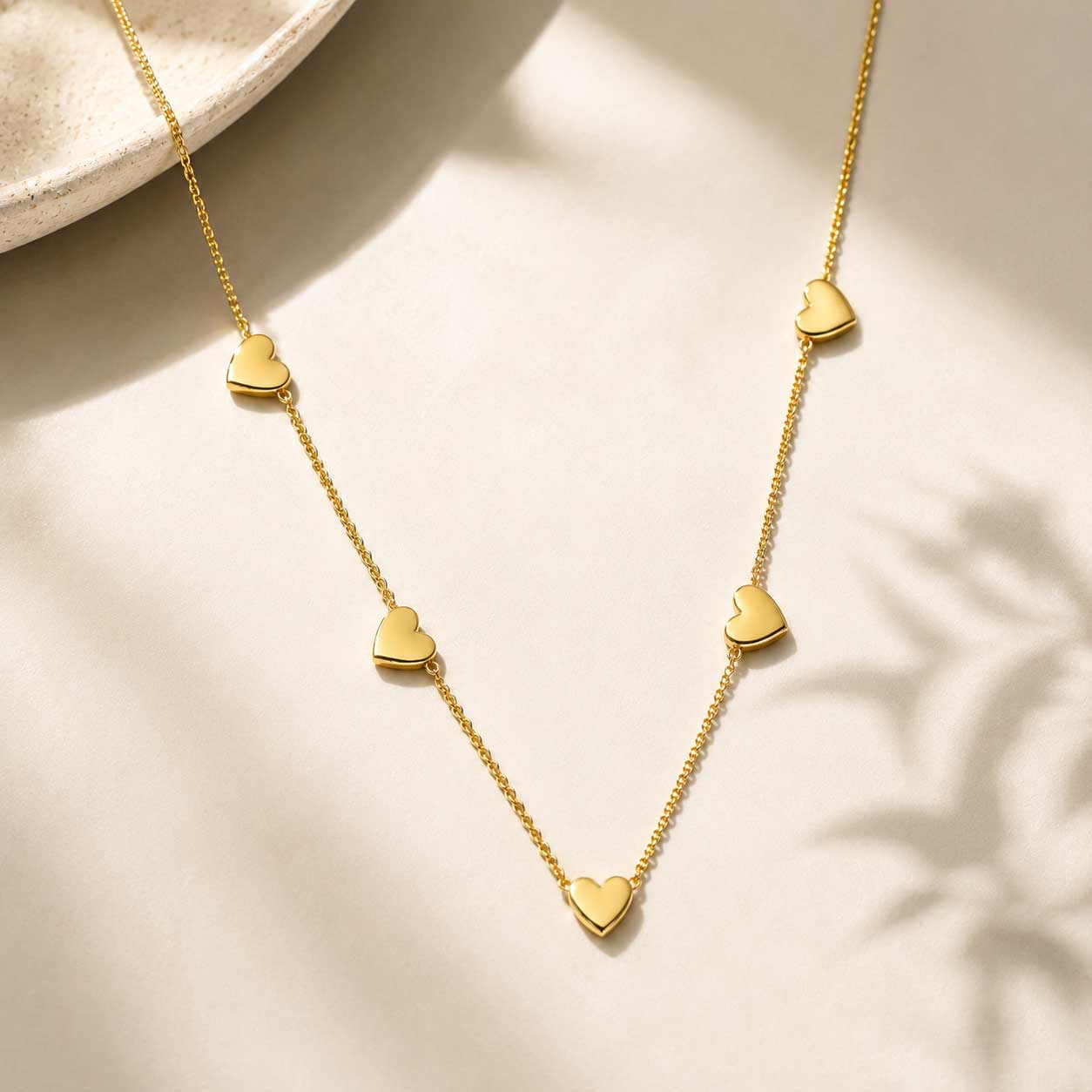 Goldene Halskette mit Herzanhängern – Golden Heart Halskette von Saltyblush, filigrane Goldkette mit 5 polierten Herzcharms, Flatlay auf hellem Hintergrund
