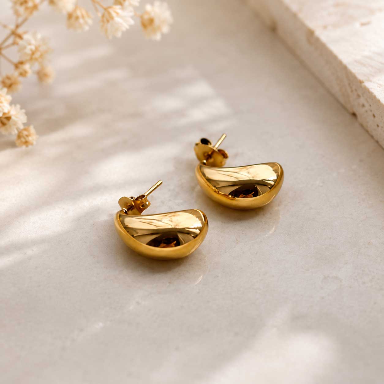 Goldene Halbmond Ohrringe – Golden Moon Ohrringe von Saltyblush, glänzende Stecker in Halbmond-Form aus Edelstahl, Flatlay auf weißem Tablett mit Trockenblumen
