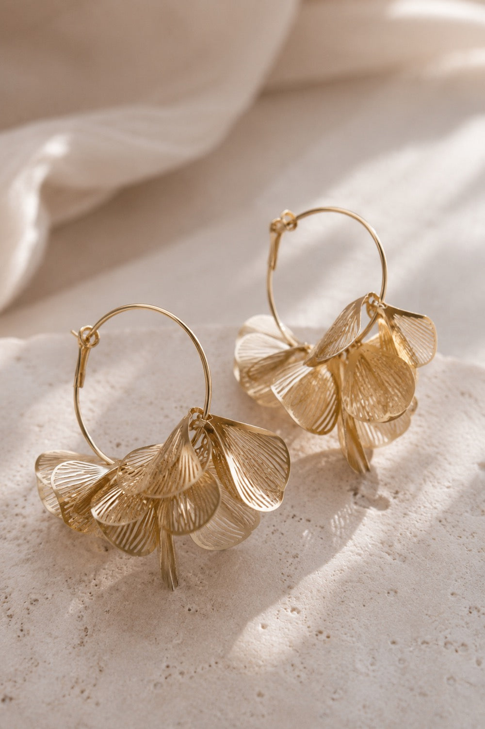 Goldene Creolen mit mehrlagigen Blütenblatt-Anhängern auf hellem Steinuntergrund – Golden Petal Hoops von Saltyblush
