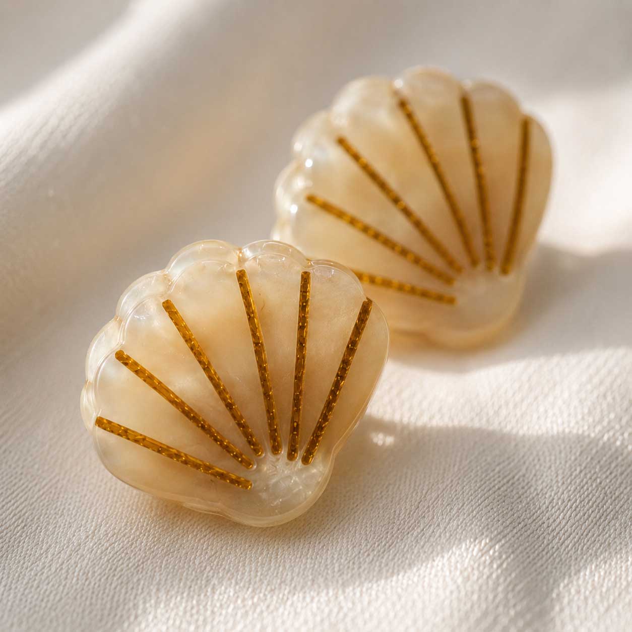 Nahaufnahme zweier Golden Shell Haarklammern in Muschelform aus cremefarbenem Acryl mit goldenen Akzenten im Sonnenlicht – Saltyblush
