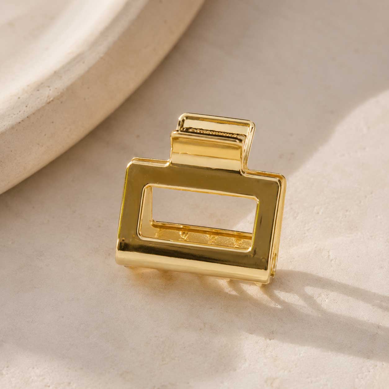 Goldene rechteckige Haarklammer auf cremefarbenem Stein im Sonnenlicht, vergoldete Metall-Haarspange mit offenem Rechteck-Design und glänzender Oberfläche
