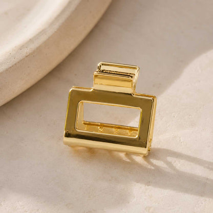 Goldene rechteckige Haarklammer auf cremefarbenem Stein im Sonnenlicht, vergoldete Metall-Haarspange mit offenem Rechteck-Design und glänzender Oberfläche
