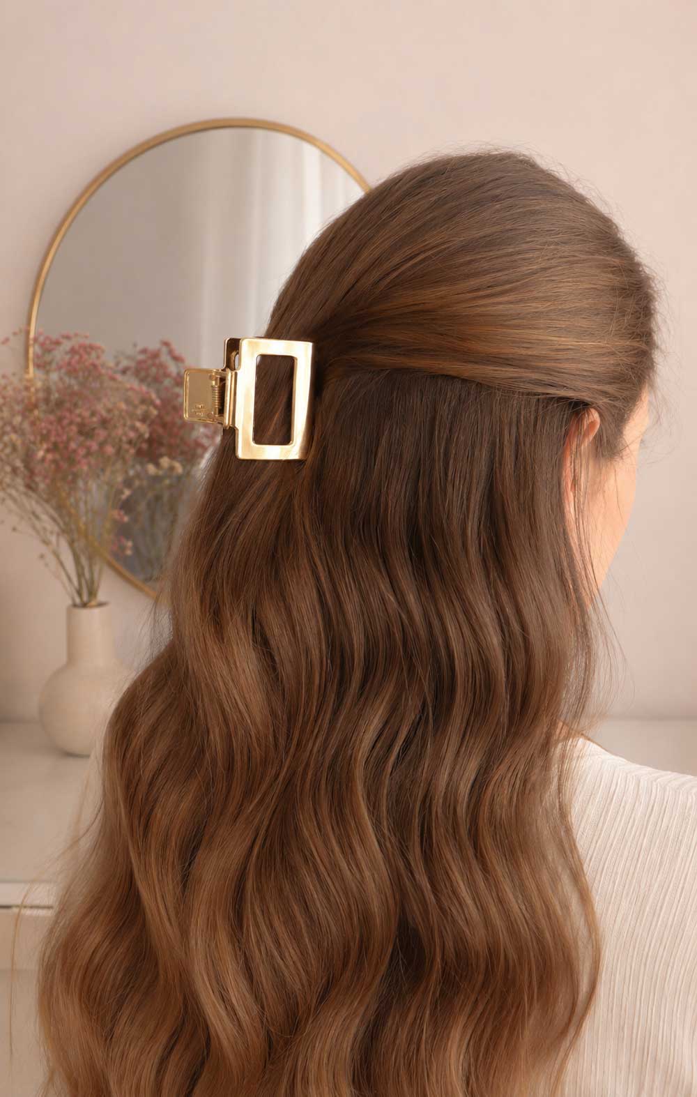 Goldene rechteckige Haarklammer im braunen Wellenhaar getragen, vergoldete Metall-Haarspange mit offenem Rechteck-Design als Half-Up-Hairstyle mit rundem Spiegel im Hintergrund
