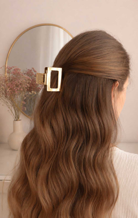 Goldene rechteckige Haarklammer im braunen Wellenhaar getragen, vergoldete Metall-Haarspange mit offenem Rechteck-Design als Half-Up-Hairstyle mit rundem Spiegel im Hintergrund
