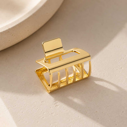 Goldene rechteckige Haarklammer Seitenansicht auf cremefarbenem Stein im Sonnenlicht, vergoldete Metall-Haarspange mit offenem Gitter-Rechteck-Design und Schattenspiel
