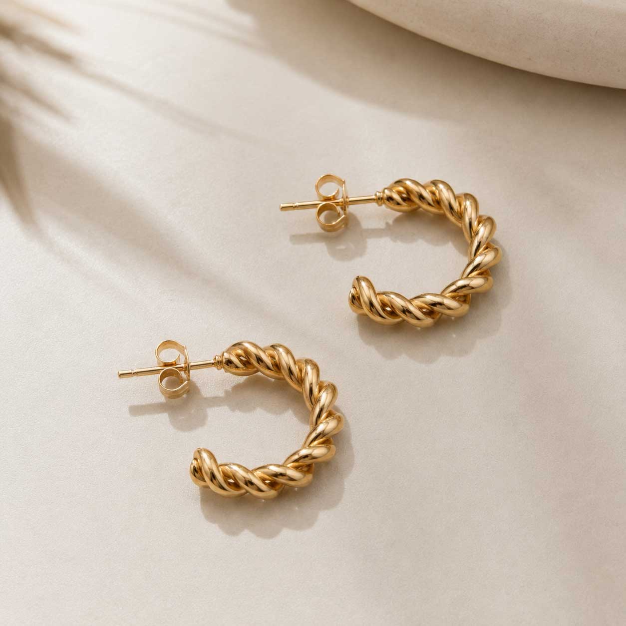 Zwei goldene Twist Creolen aus Edelstahl mit gedrehtem Design auf cremefarbenem Stoff – Saltyblush
