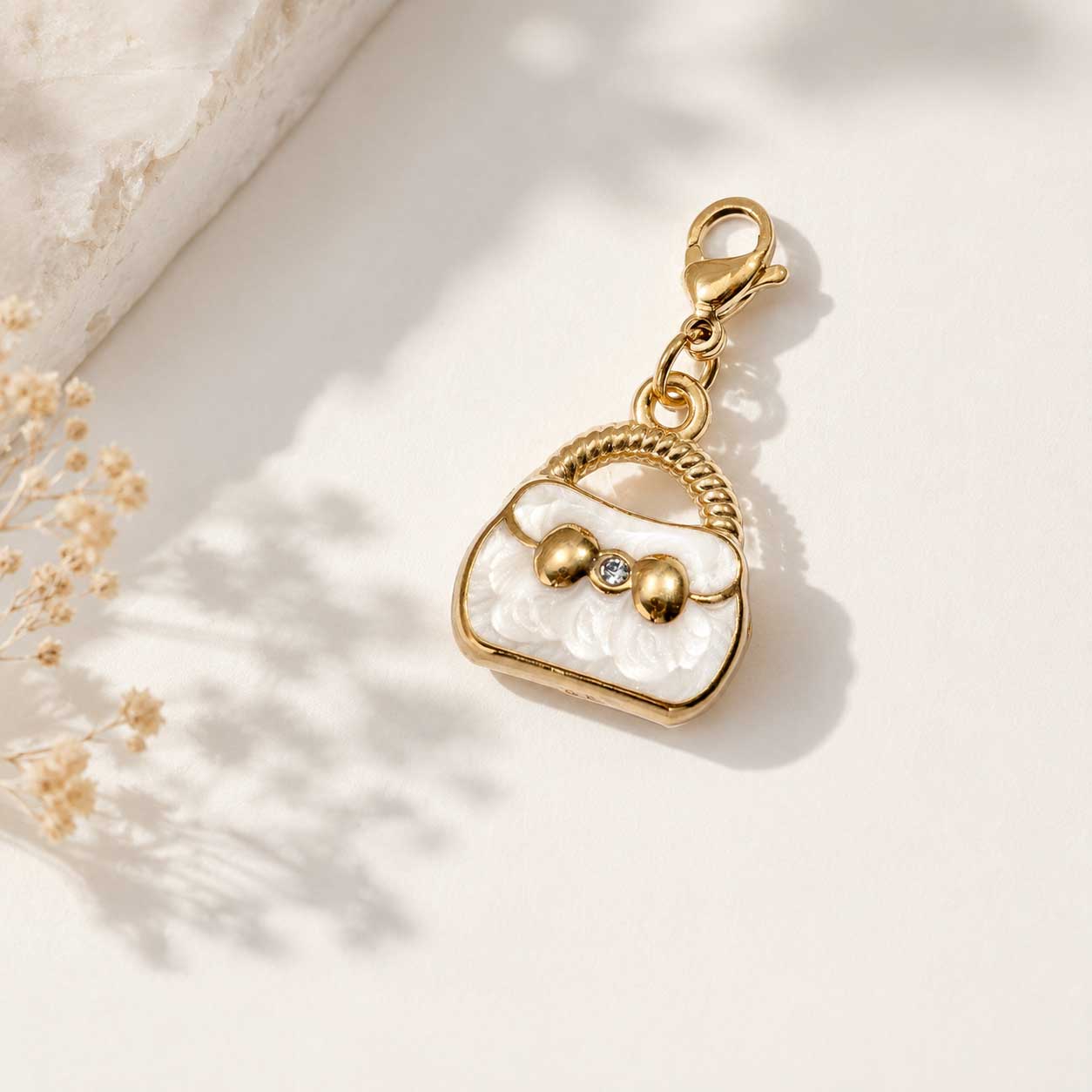 Goldener Handtaschen-Charm in Weiß mit Kristallstein und Karabinerverschluss auf cremefarbenem Hintergrund mit Trockenblumen – Saltyblush
