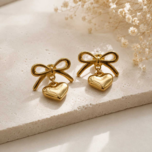Zwei goldene Heart Bow Ohrringe mit Schleife und Herzanhänger aus Edelstahl auf cremefarbenem Steinuntergrund mit Trockenblumen – Saltyblush
