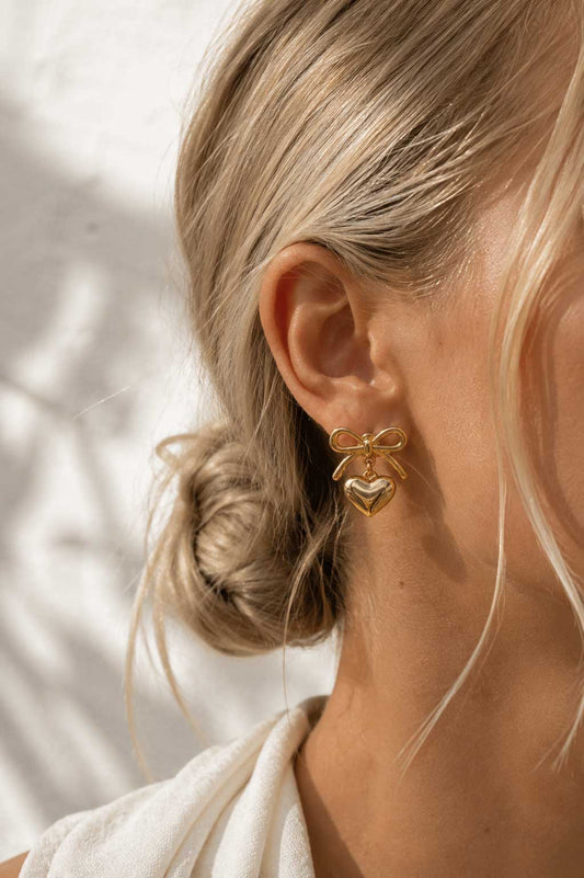 Blonde Frau mit hochgestecktem Haar trägt goldene Heart Bow Ohrringe aus Edelstahl – elegante Lifestyle-Aufnahme – Saltyblush
