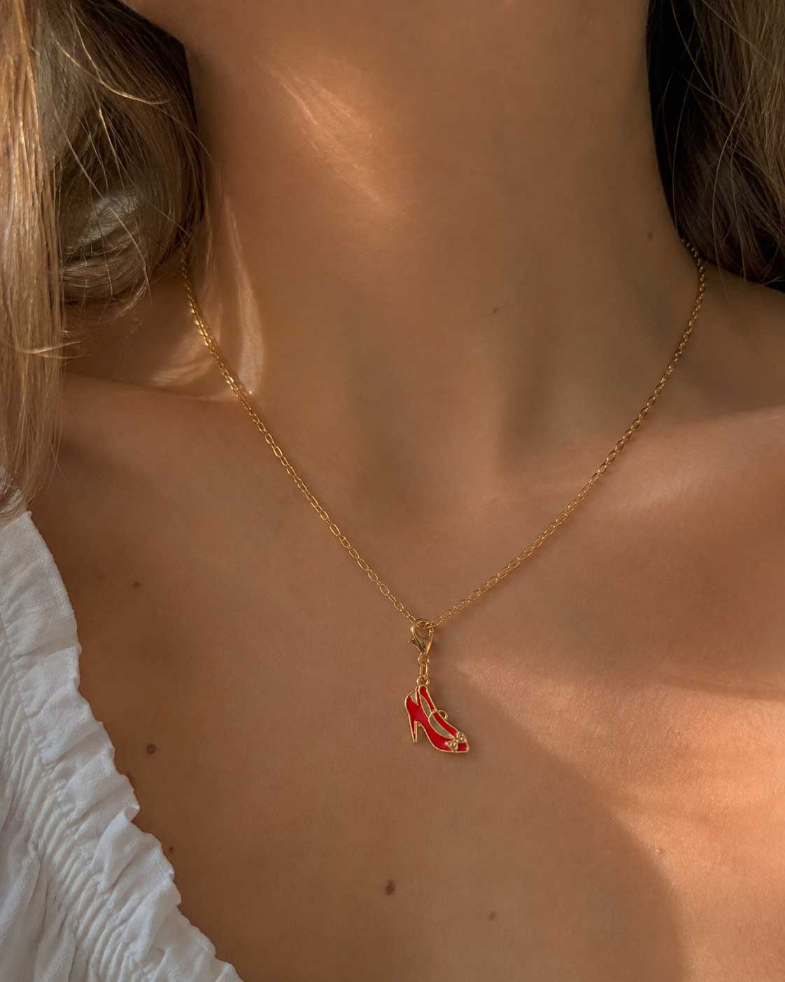 Frau in weißem Rüschenoberteil trägt goldene Halskette mit rotem High Heel Charm am Décolleté im Sonnenlicht – Saltyblush
