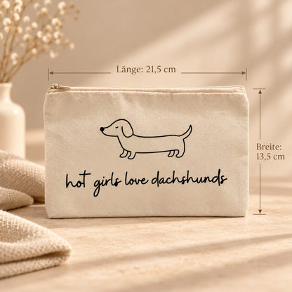 Beige Kosmetiktasche mit schwarzer Dackelzeichnung, Schriftzug „hot girls love dachshunds“ und Reißverschluss. Maßangaben: 21,5 cm Länge und 13,5 cm Breite.
