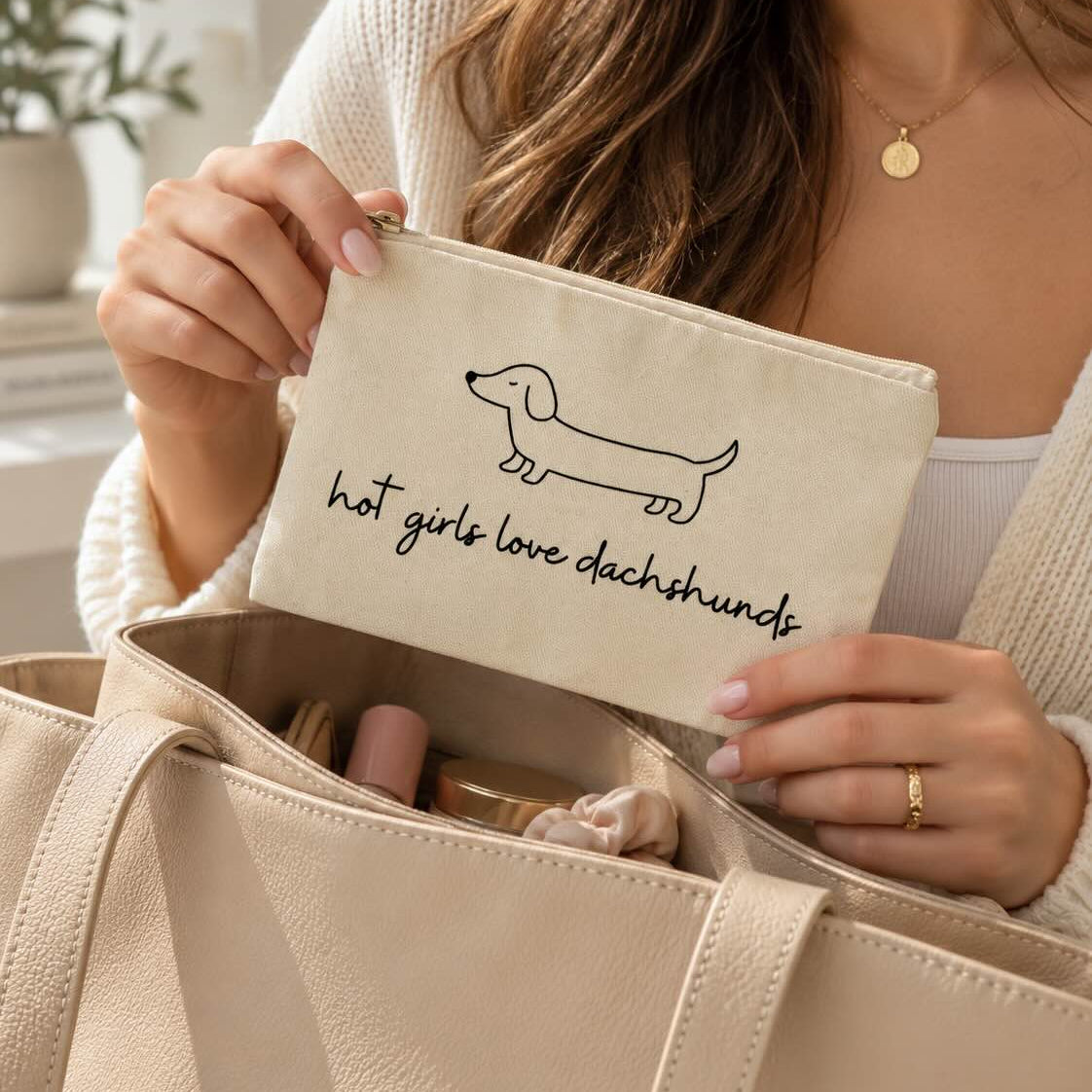Frau hält die beige Kosmetiktasche mit Dackelmotiv und Schriftzug „hot girls love dachshunds“ über einer geöffneten Handtasche mit Beauty-Produkten.
[AI]