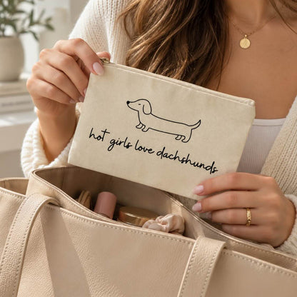 Frau hält die beige Kosmetiktasche mit Dackelmotiv und Schriftzug „hot girls love dachshunds“ über einer geöffneten Handtasche mit Beauty-Produkten.
[AI]