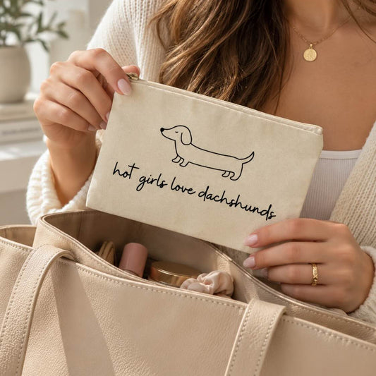 Frau hält die beige Kosmetiktasche mit Dackelmotiv und Schriftzug „hot girls love dachshunds“ über einer geöffneten Handtasche mit Beauty-Produkten.
[AI]