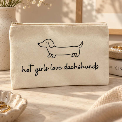 Beige Kosmetiktasche mit Reißverschluss, schwarzer Dackelzeichnung und Schriftzug „hot girls love dachshunds“ neben einem Handtuch im warmen Sonnenlicht.

