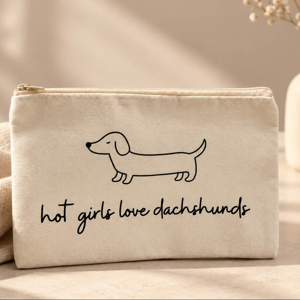 Beige Kosmetiktasche mit schwarzem Dackelmotiv und Schriftzug „hot girls love dachshunds“ auf einem hellen Tisch neben einer Schmuckschale und weißen Blumen.
