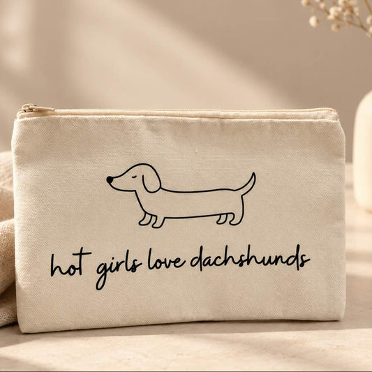 Beige Kosmetiktasche mit schwarzem Dackelmotiv und Schriftzug „hot girls love dachshunds“ auf einem hellen Tisch neben einer Schmuckschale und weißen Blumen.
