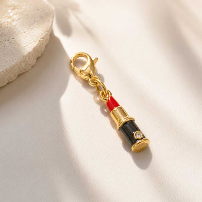 Goldener Lippenstift-Charm mit roter Emaille und schwarzem Gehäuse auf cremefarbenem Untergrund, vergoldeter Edelstahl-Charm mit Karabinerverschluss und Zirkonia-Stein
