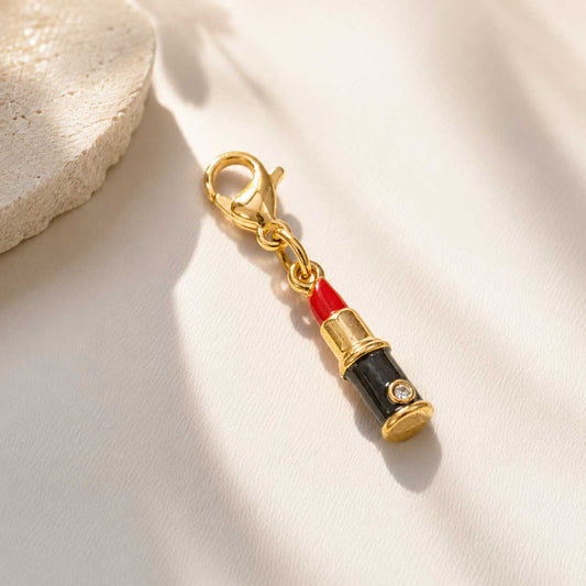 Goldener Lippenstift-Charm mit roter Emaille und schwarzem Gehäuse auf cremefarbenem Untergrund, vergoldeter Edelstahl-Charm mit Karabinerverschluss und Zirkonia-Stein
