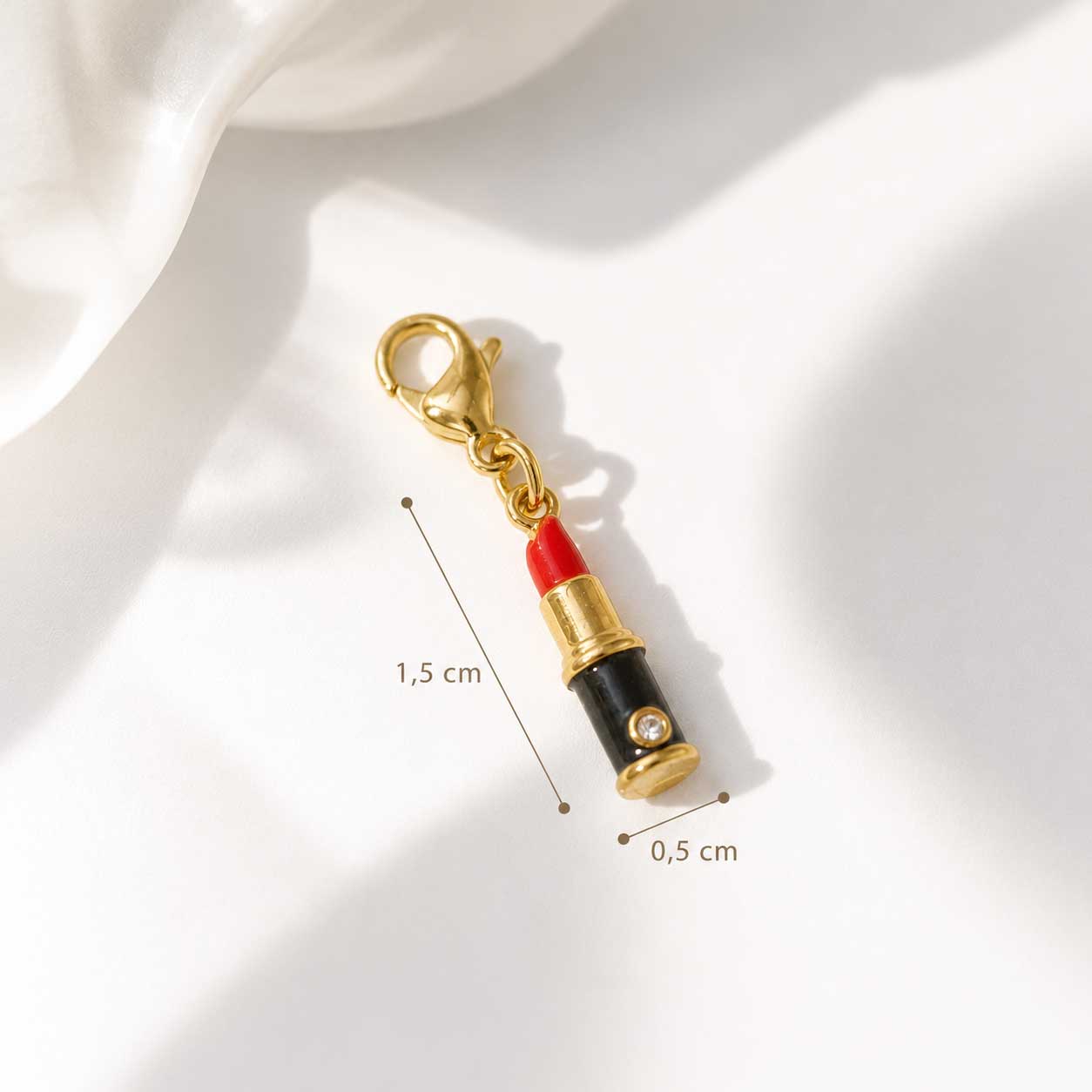 Lippenstift-Charm Infografik – goldener Emaille-Charm Höhe 1,5 cm, Breite 0,5 cm, roter Lippenstift mit schwarzem Gehäuse und Karabinerverschluss auf weißem Seidenstoff
