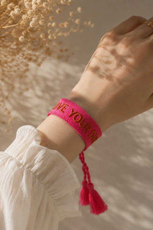 Pinkes Live Your Dreams Armband mit Quasten am Handgelenk getragen, weißes Kleid, Trockenblumen im Hintergrund – Saltyblush
