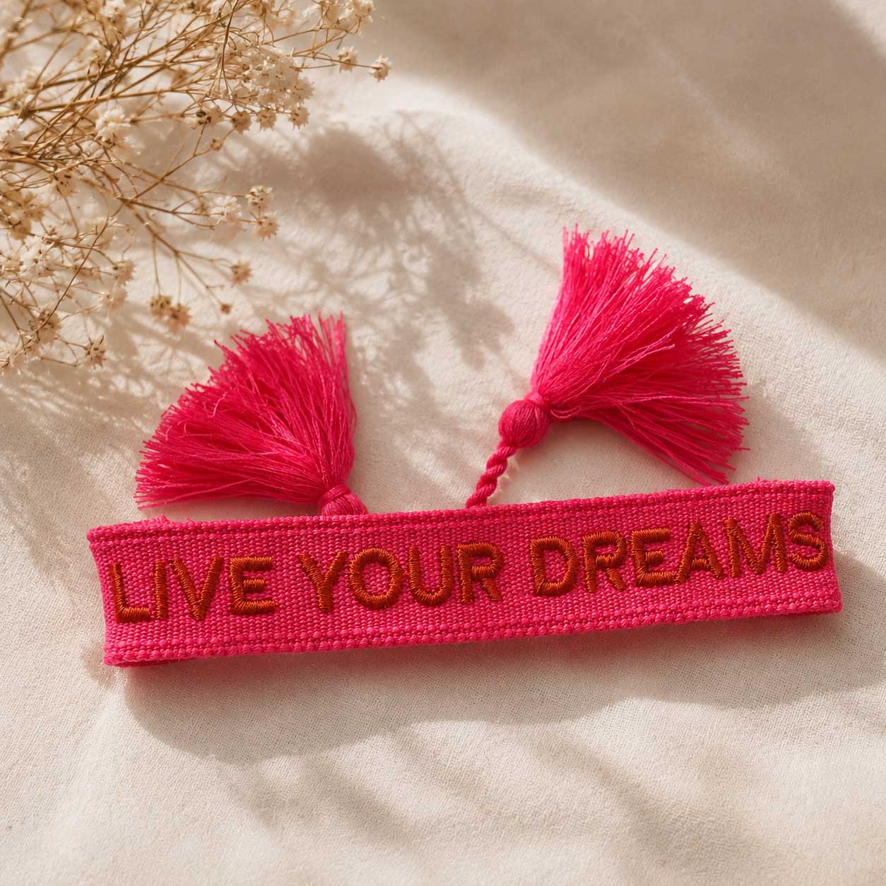 Pinkes Statement Armband Live Your Dreams mit zwei pinken Quasten auf hellem Stoff, Draufsicht – Saltyblush
