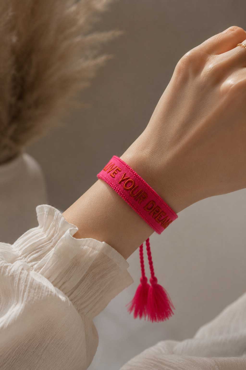 Pinkes Live Your Dreams Armband mit Quasten am Handgelenk einer Frau in weißer Bluse – Saltyblush
