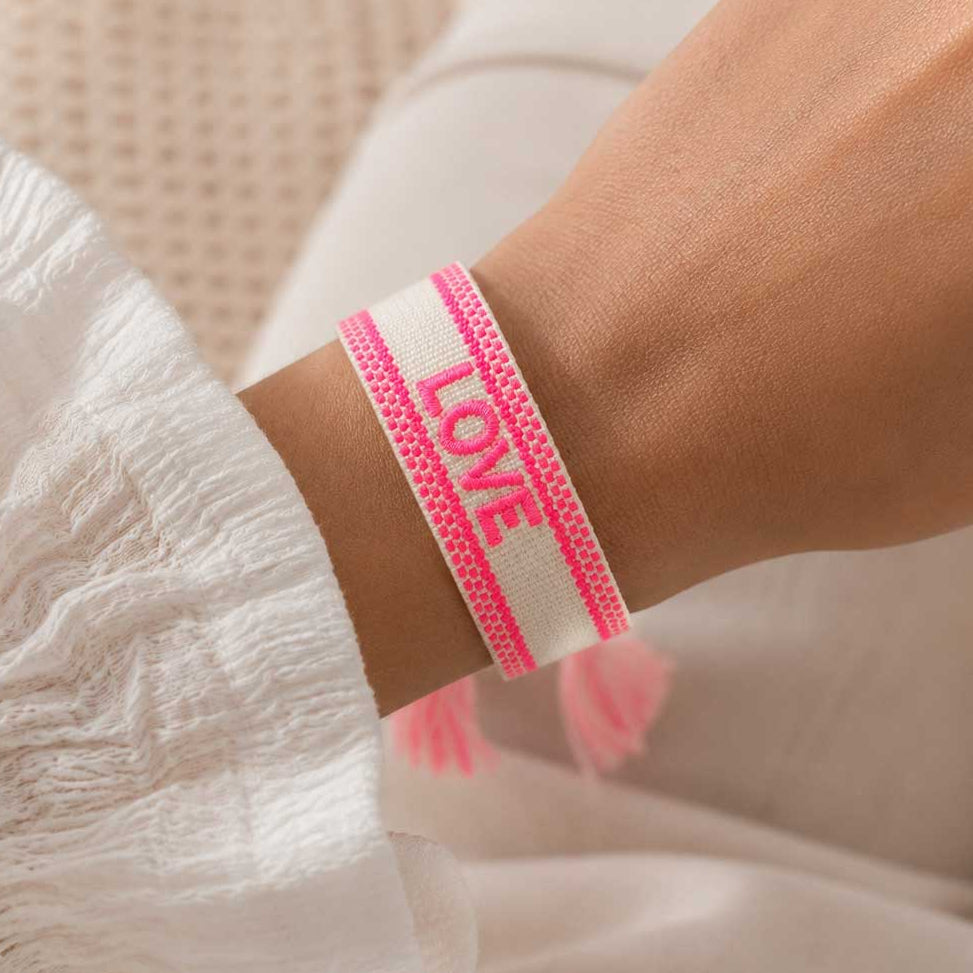 Neonpinkes LOVE Armband am Handgelenk getragen, gewebtes Stoffarmband mit pinken Streifen zu weißem Spitzenoberteil
