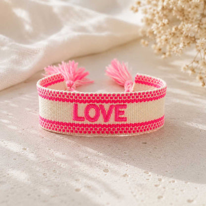 Cremefarbenes gewebtes LOVE Armband mit neonpinken Streifen und rosa Quasten auf hellem Untergrund mit Schleierkraut
