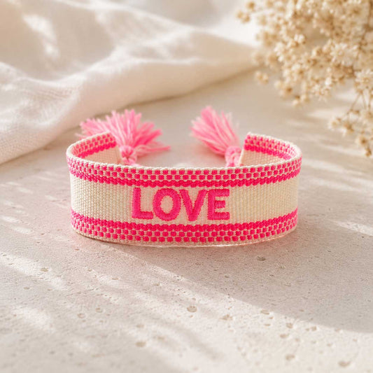 Cremefarbenes gewebtes LOVE Armband mit neonpinken Streifen und rosa Quasten auf hellem Untergrund mit Schleierkraut
